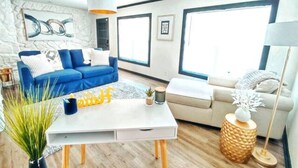 A modern living room with a blue sofa, a white coffee table, and a beige sofa. There are large windows, a stone wall, and a hanging light fixture. - Maison Modernè Unit A: Work + Play (San Antonio)