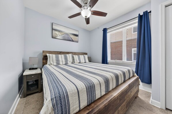 A bedroom with a bed, a nightstand, a ceiling fan, a window with curtains, and a wall-mounted picture.