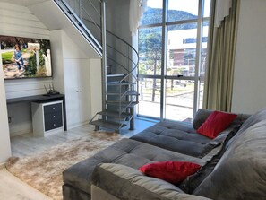 Uma sala de estar moderna com um sofá cinzento, uma pequena escrivaninha, uma escada e uma janela grande com vista para a rua e colinas.