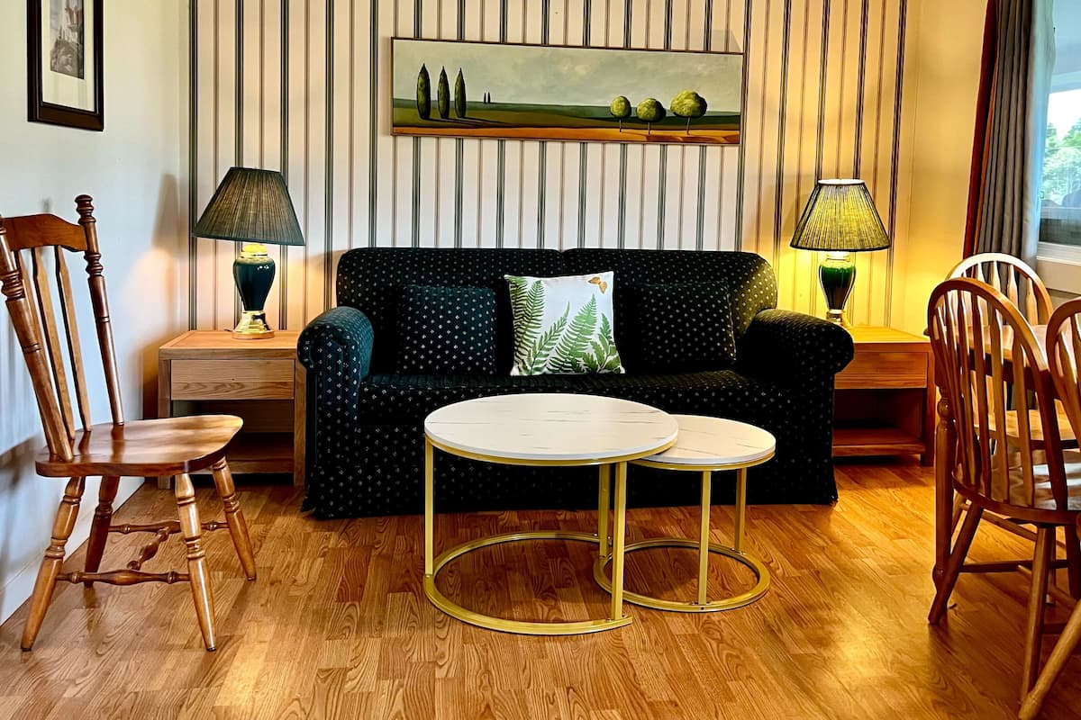 A living room with a striped wall, a painting, a sofa, two lamps, a table, and wooden chairs.