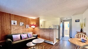A cozy living room with a wooden wall, a sofa with cushions, a coffee table, and a dining table with chairs. There is a kitchen area visible through an open doorway. - St Lawrence Motel (North Rustico)