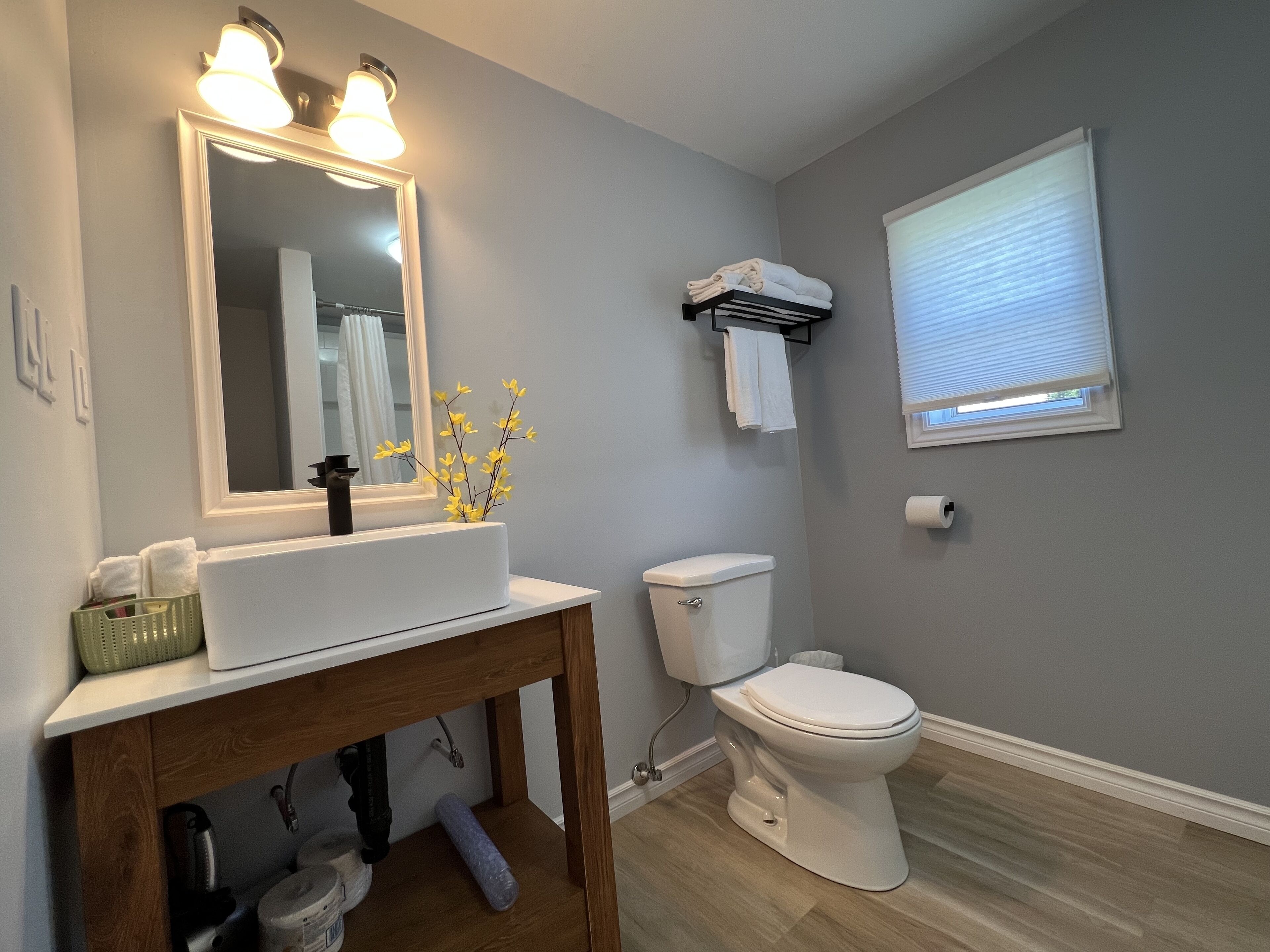 A bathroom with a white sink, mirror, and toilet. There is a window with blinds, a towel rack, and a small plant on the sink.