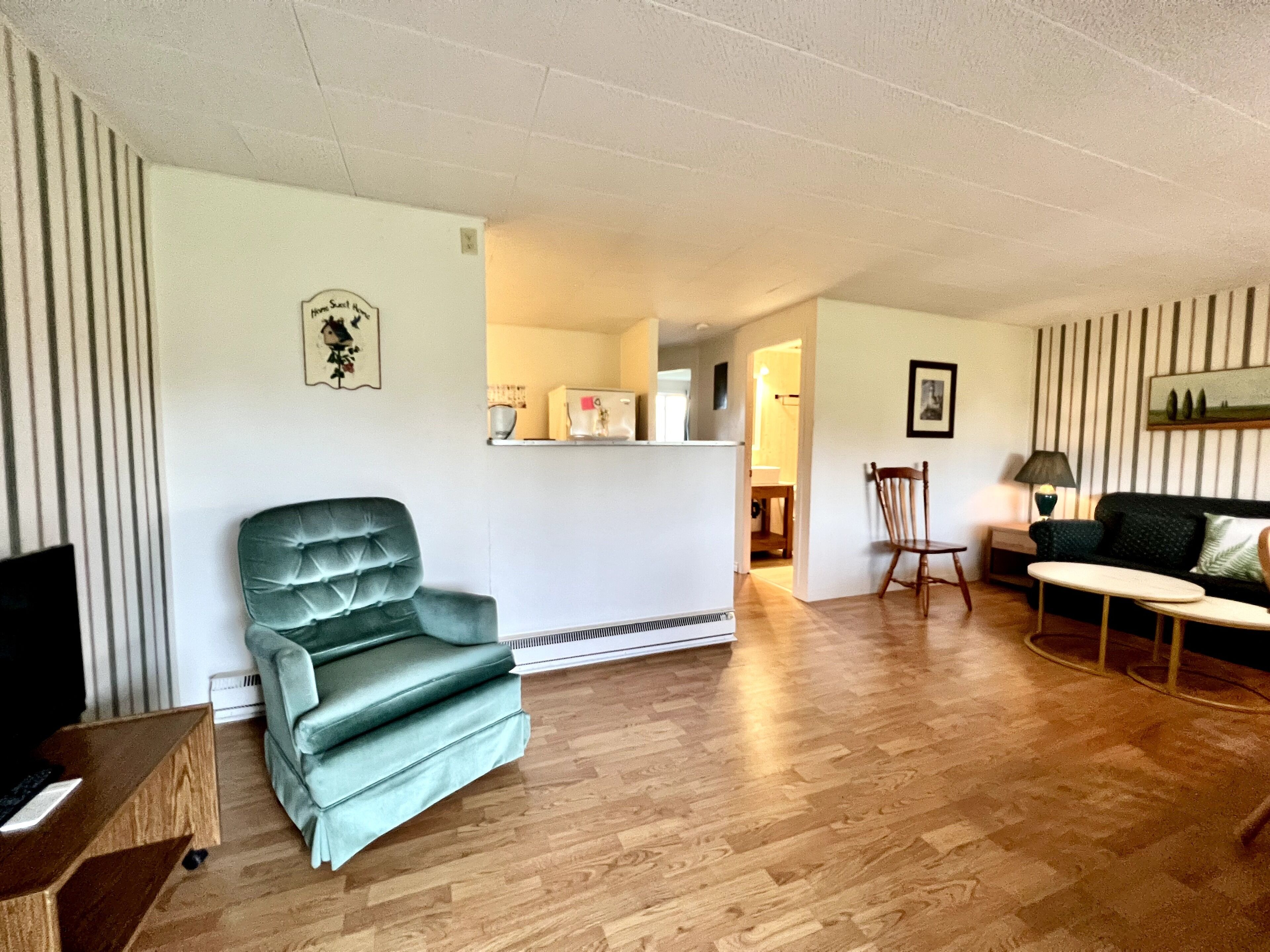 A living room with a green armchair, a wooden coffee table, and a sofa. There is a kitchen area in the background with a refrigerator and a microwave.
