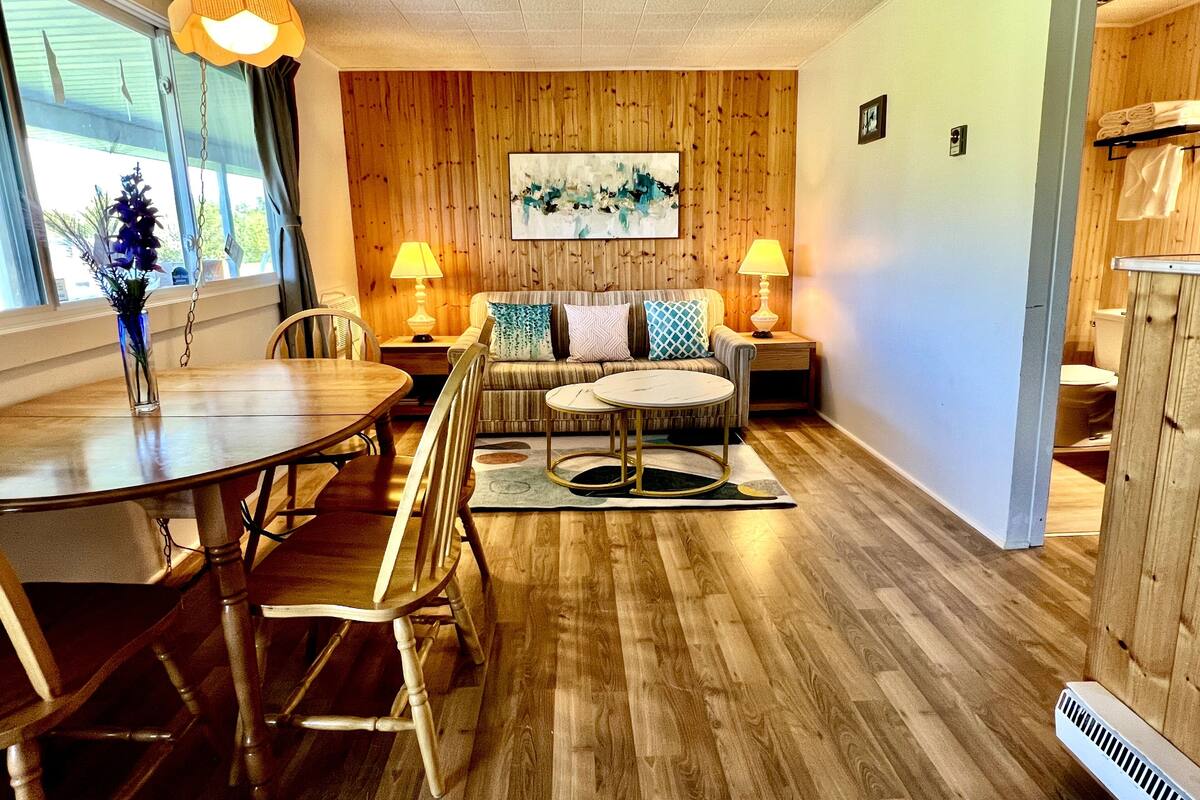 A cozy living room with a wooden wall, a round coffee table, a sofa with patterned cushions, and a dining table with chairs.