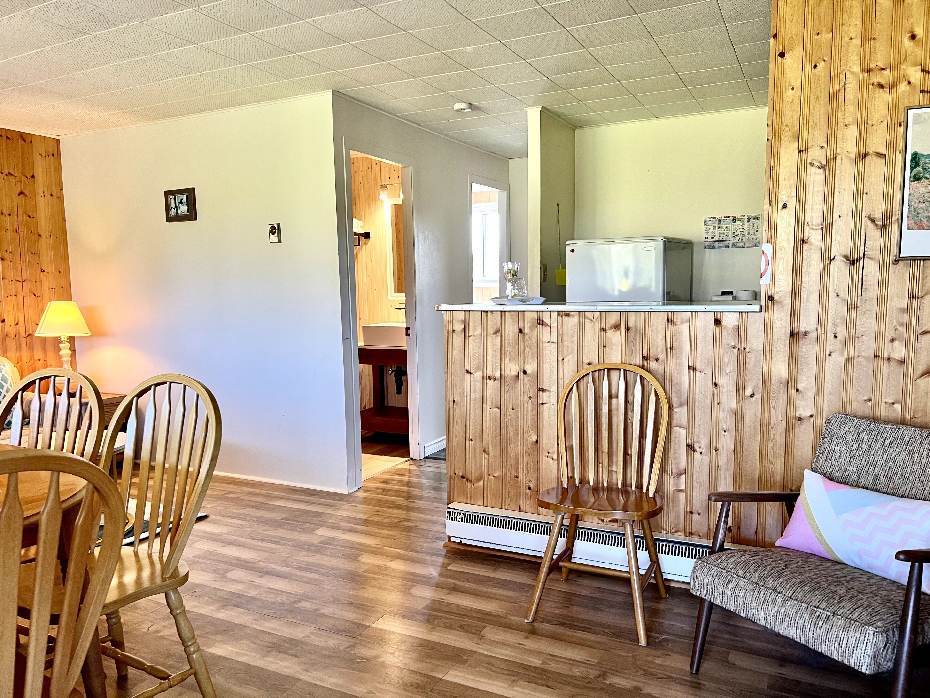 A room with wooden flooring, a dining table with chairs, a sofa, a kitchen area with a refrigerator, and a wooden wall panel.