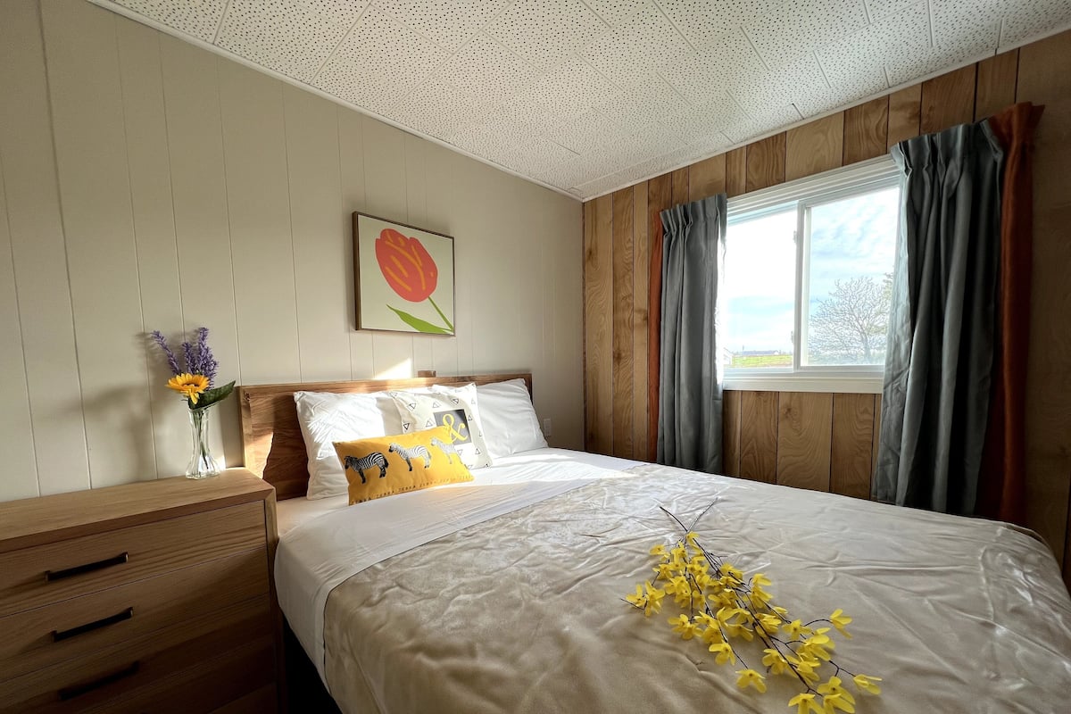 A bedroom with a wooden headboard, a bed with white bedding, a nightstand with a vase of flowers, and a window with curtains.