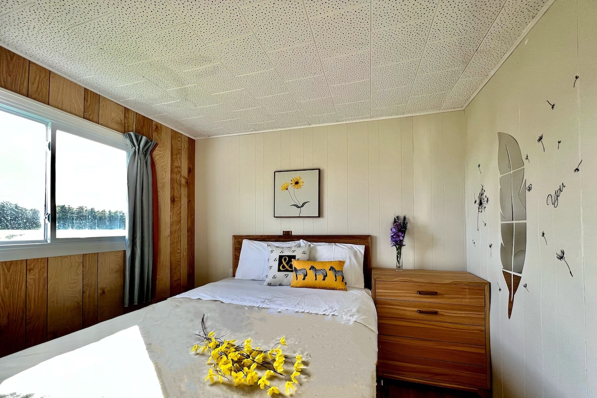 A bedroom with a wooden headboard, a bed with white bedding and decorative pillows, a nightstand with a vase of flowers, and a window with curtains.