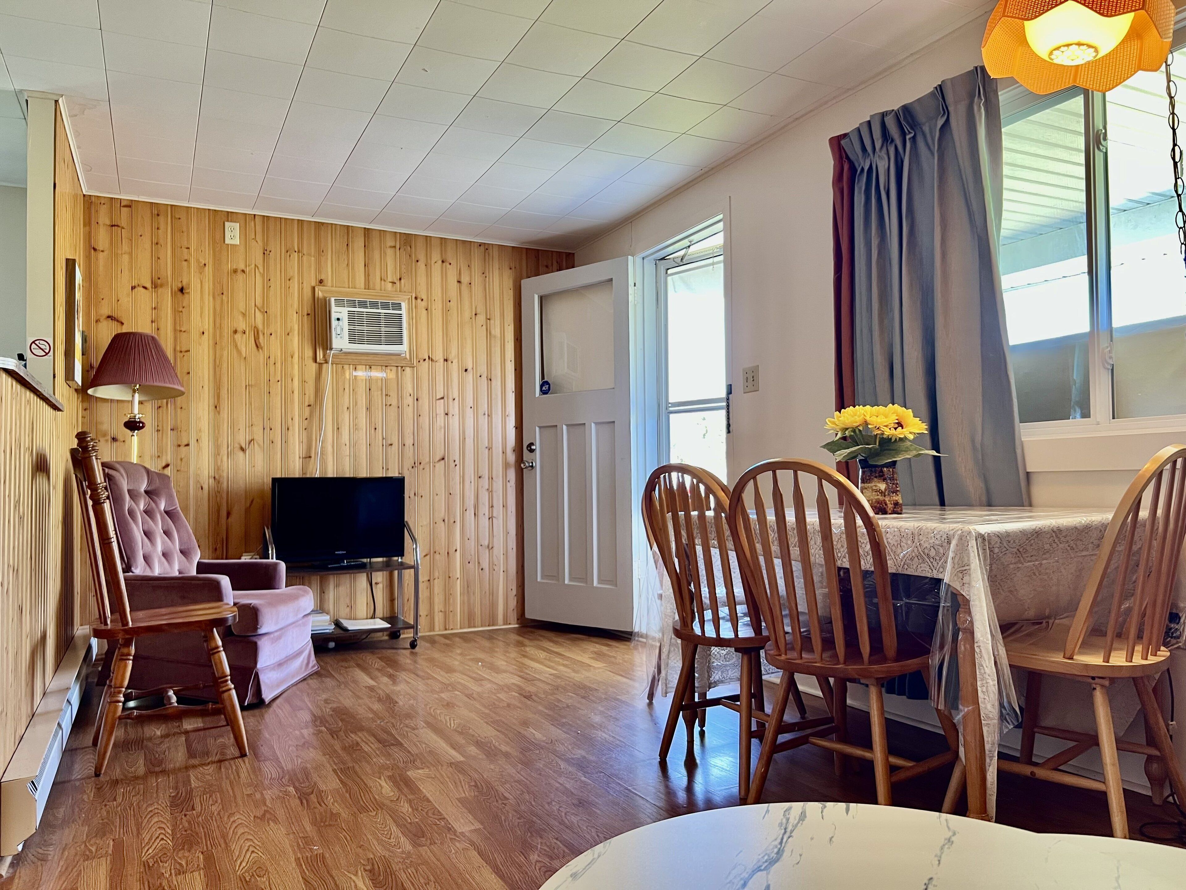 A room with wooden panel walls, a television on a stand, a dining table with chairs, and a window with curtains.