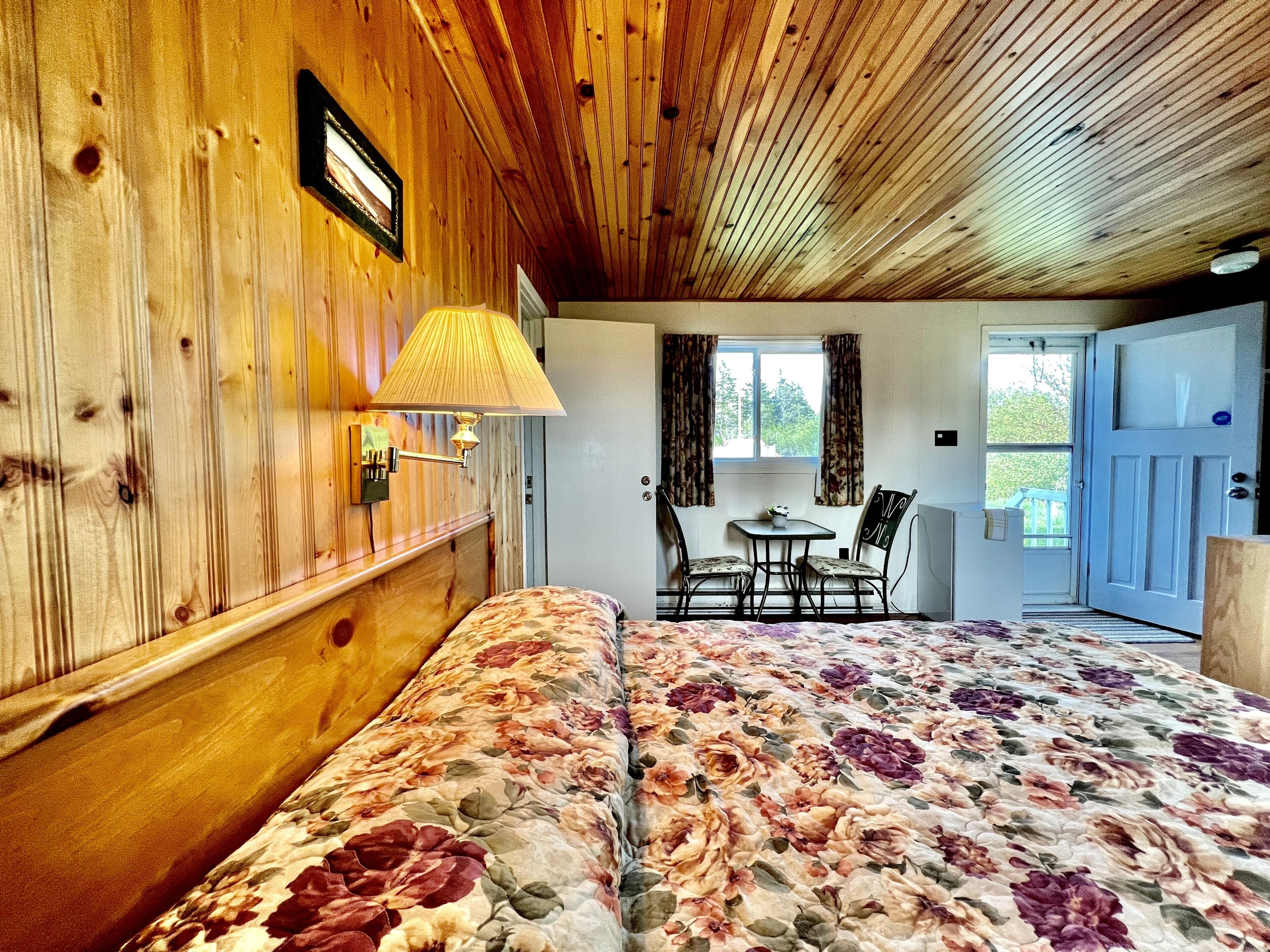 Habitació amb revestiment de fusta, un llit amb roba de llit floral, una taula amb cadires i una finestra amb cortines.