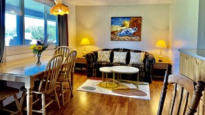 A living room with a dining area, wooden furniture, a painting on the wall, and a window with curtains. - St Lawrence Motel (North Rustico)