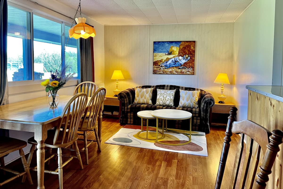 A living room with a dining area, wooden furniture, a painting on the wall, and a window with curtains.