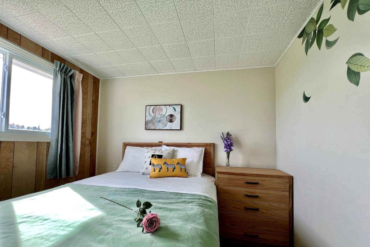 A bedroom with a wooden headboard, a bed with a green bedspread, a single pink rose, a wall-mounted picture, and a nightstand with a vase of purple flowers.