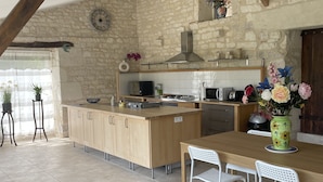 Una cocina con pared de piedra, vigas de madera, una isla central y un comedor con sillas blancas.