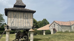 Una construcción de piedra con techo cónico, posiblemente un edificio histórico, en un área cubierta de césped con una casa de piedra al fondo.