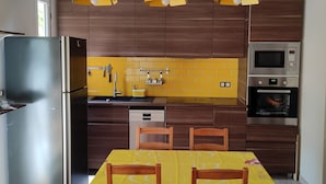 A modern kitchen with wooden cabinets, a stainless steel refrigerator, a microwave, and an oven. There is a dining table with chairs and yellow pendant lights above. - Renovated apartment (Marseille)