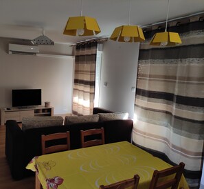 A living room with a sofa, a TV, a dining table with chairs, and yellow pendant lights.
