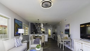A modern living room with a sofa, coffee table, and TV. There is a dining area with chairs and a table. The room has a kitchen in the background. - Spectacular Direct Oceanfront Condo ~ Wraparound Balcony  ~ Top Floor! (Saint Augustine)