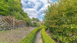 A stone wall with a wooden gate, a gravel path, and lush green foliage. - Bowmanstead Cottage - quirky & cosy traditional village cottage (Coniston)