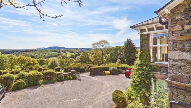 A stone building with a gravel driveway leading to a red car parked near a well-maintained garden with various shrubs and a yellow bench. - Drawing Room, grand apartment with stunning lake views across Beatrix Potter country (Far Sawrey)