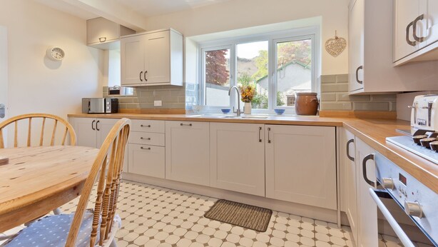 A modern kitchen with white cabinets, a wooden dining table, and a window with a view of trees. - Green Croft - garden cottage in Coniston village (CONISTON)