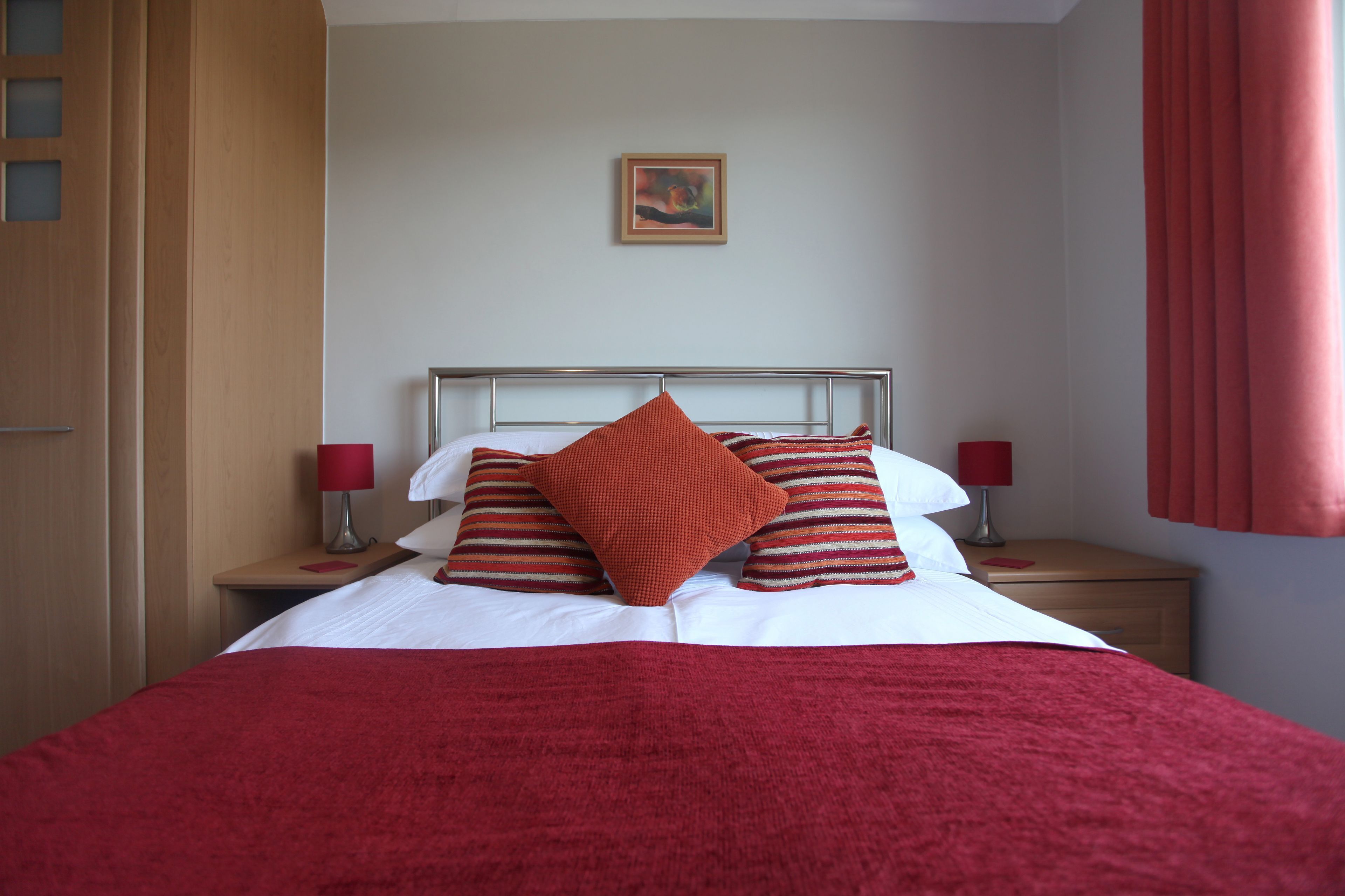 A neatly made bed with a red blanket and pillows, a framed picture on the wall, and a wooden nightstand with a lamp.