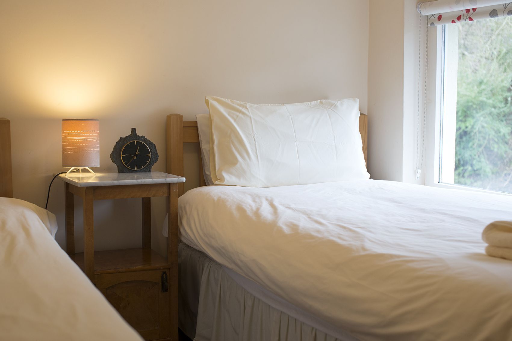 A bedroom with a bed, a bedside table with a lamp, and a window with a view of greenery.