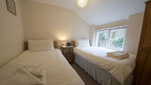 A bedroom with two single beds, a bedside table with a lamp, a window with a view of greenery, and a wooden wardrobe. - Poppy Cottage, village cottage with lake views (Coniston)