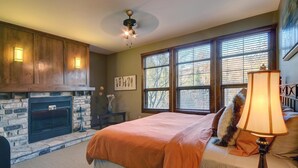 A bedroom with a stone fireplace, a bed with an orange blanket, a lamp, and a window with blinds. - Elegant 4BR Condo for 8 • Ski Views • Walk to Village • Spa & Shuttle (Mont-Tremblant)