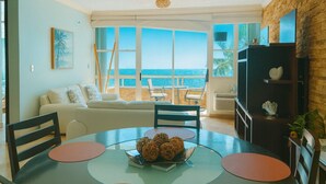 A living room with a large window overlooking the ocean, a sofa, a dining table with chairs, and a TV stand with decorative items. - Pelican Reef 507 Beachfront 2bdr apt (Rincón)