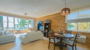 A spacious living room with a large window, a fireplace, and a dining area with a round table and chairs. - Pelican Reef 507 Beachfront 2bdr apt (Rincón)