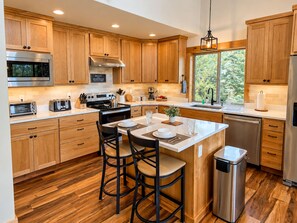A modern kitchen with wooden cabinets, stainless steel appliances, a central island with bar stools, and a view through the window.