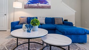 Ein Wohnzimmer mit einem blauen Ecksofa, zwei runden Tischen, einer Topfpflanze und einem Gemälde von Baum und Mond.