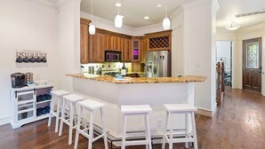A modern kitchen with a central island, white bar stools, wooden cabinets, and a stainless steel refrigerator. - Huge Home w/ Gameroom, Patio, & Secret Room! (Dallas)