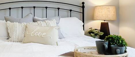 Una cama bien tendida con ropa blanca y almohadas decorativas, una lámpara de noche y una bandeja con una planta en maceta.