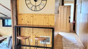 Una zona de bar con barra de madera, un reloj de pared grande, dos taburetes y un revestimiento de pared de madera.