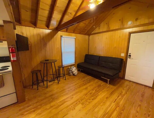 Ein gemütliches Blockhaus mit Holzwänden und geneigtem Dach. Es verfügt über eine Küchenzeile, ein Sofa, einen kleinen Tisch und ein Fenster mit Jalousien.