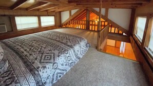 A bedroom with a large bed, a wooden ceiling, and a view of the lower level through a window.