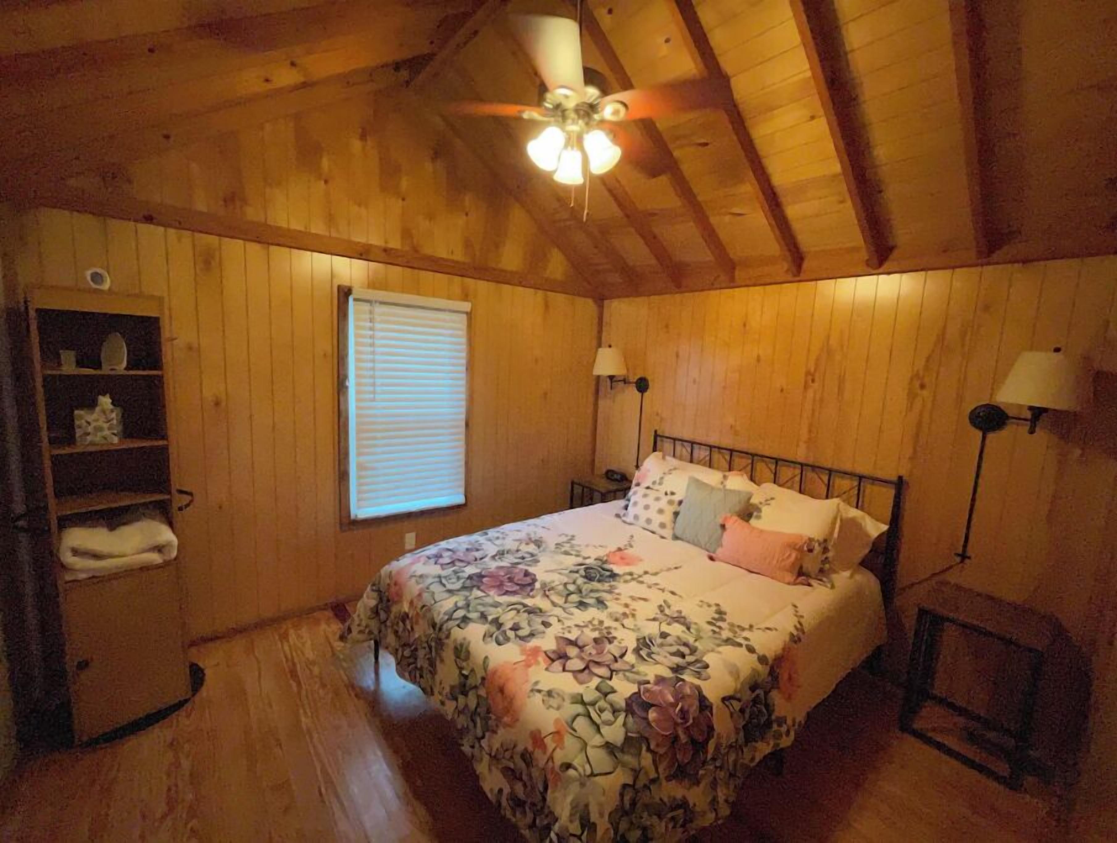 A bedroom with wooden walls, a bed with floral bedding, a ceiling fan, a window with blinds, and a nightstand with a lamp.