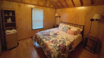 A bedroom with wooden walls, a bed with floral bedding, a ceiling fan, a window with blinds, and a nightstand with a lamp.