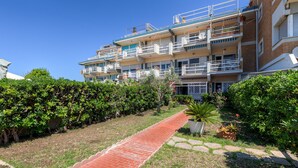 Un immeuble résidentiel à plusieurs étages, doté de balcons, d’un chemin pavé en briques et d’un espace vert bien entretenu.