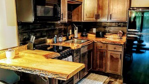 A kitchen with wooden cabinets, a black refrigerator, a microwave, and a stove. There are two bar stools and a rug on the floor. - The Oaklyn Container ~ Hocking Hills ~ Hot tub ~ Firepit ~ Wifi (Logan)