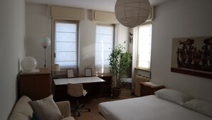 Une chambre d’hôtel avec un lit, un bureau, une chaise, une plante en pot et une fenêtre avec des rideaux.