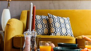 A living room with a mustard yellow sofa, patterned pillows, a decorative wall panel, a vase with dried flowers, and a table with breakfast items. - The Majestic Garden Suite (Paris)