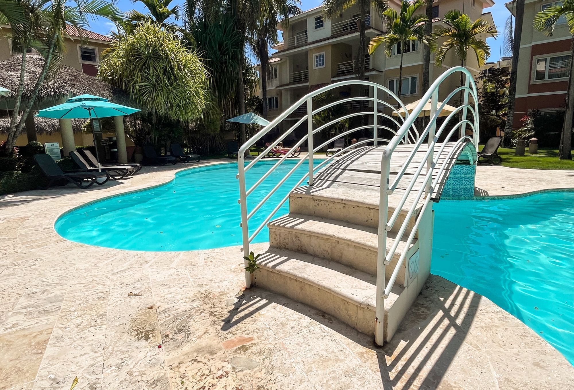 Ein Swimmingpool mit einer Treppe zu einer Sonnenterrasse, Liegestühlen, Sonnenschirmen und Palmen.