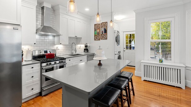 A modern kitchen with white cabinetry, stainless steel appliances, a large island with bar stools, pendant lighting, and a view of the outdoors through a window.