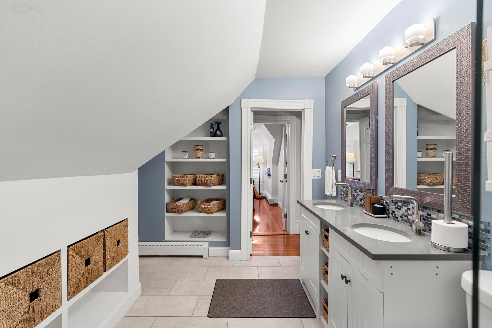 A modern bathroom with a large mirror, two sinks, and wicker baskets on shelves.