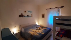 A bedroom with a bunk bed, a bed with a patterned bedspread, a nightstand, a wall-mounted shelf with a painting, and a window with curtains. - Castiglioncello Baia del Quercetano, the sea in the eyes. (Rosignano Solvay-Castiglioncello)