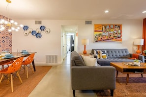 Un salon moderne avec un canapé gris, des fauteuils orange et un coin repas comprenant une table et des chaises. Un tableau coloré orne le mur, et un luminaire est fixé au plafond.