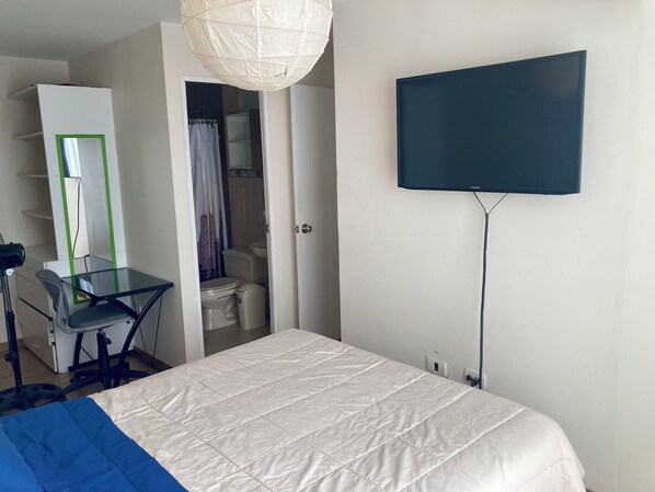 A bedroom with a bed, a desk with a chair, a television, and a bathroom visible through an open door.