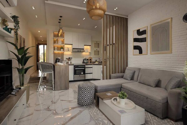 Una sala moderna con un sofá gris, una mesa de centro y un televisor. Se ve una cocina al fondo con gabinetes blancos y un lámpara colgante.